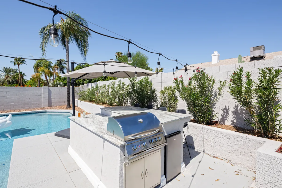 This exterior image features a sparkling swimming pool surrounded by a BBQ grill, umbrella-shaded areas, and lush landscape plantings. The scene captures the essence of outdoor summer fun and the amenities designed for group relaxation and entertaining.