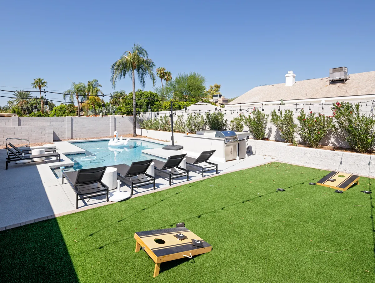An inviting scene featuring a swimming pool flanked by lounge chairs, a grill, a green lawn, and a visible cornhole game set up beside the pool. The lively composition highlights spaces for swimming, sunbathing, barbecuing, and outdoor games, creating a sense of active, social enjoyment.