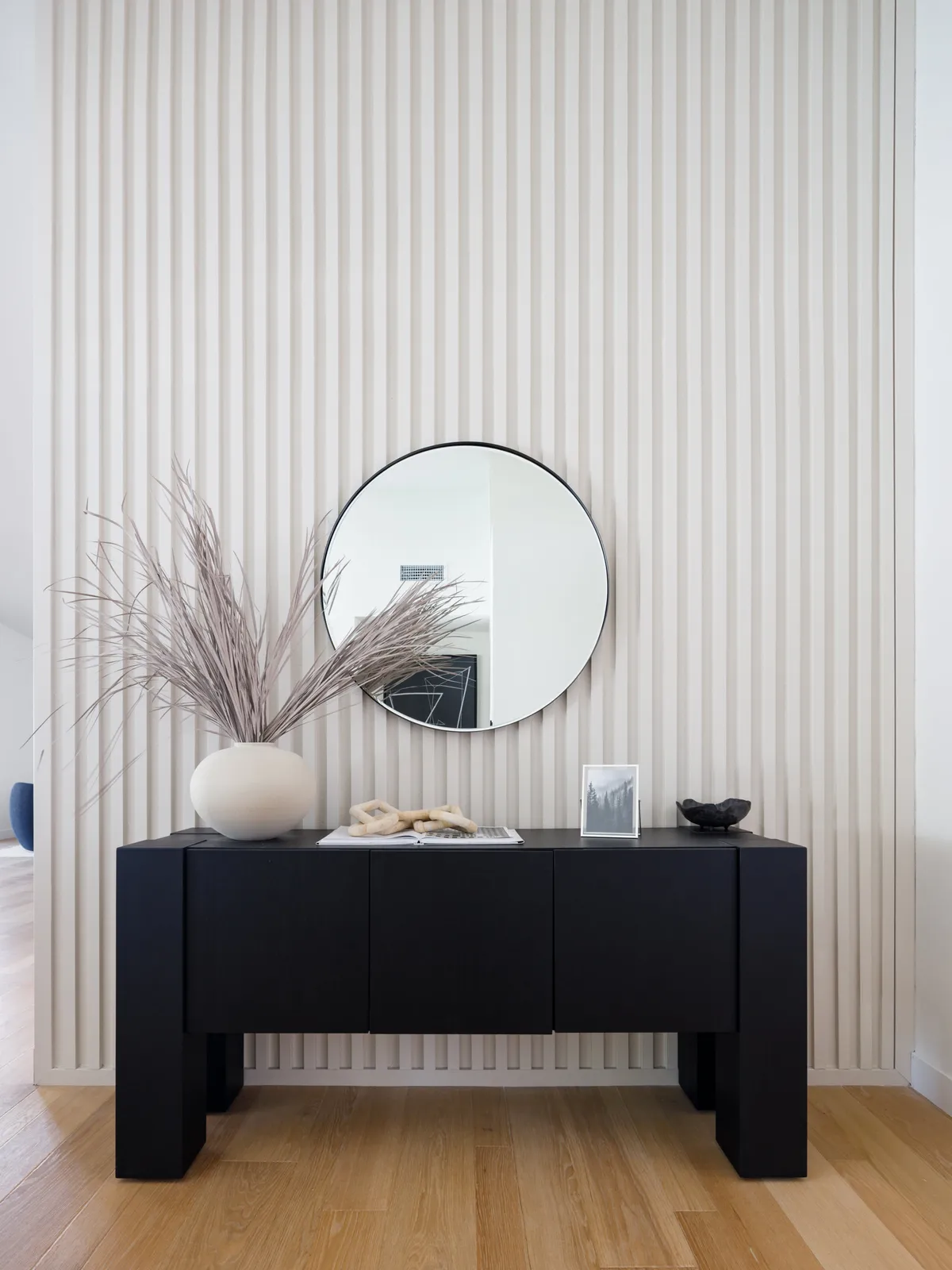 A minimalist, inviting entryway with a black console table, round mirror, a dried plant arrangement, photograph, and decorative bowl—setting a welcoming, cared-for first impression upon entering the home.