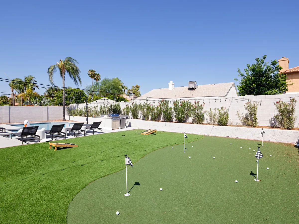 A wide view of a lush, resort-style backyard showcasing a sparkling swimming pool, jacuzzi, putting green, lounge chairs, and an outdoor grill set amidst vibrant landscaping. The scene captures guests practicing golf, sunbathing, and preparing for a barbecue, evoking an energetic yet relaxed atmosphere perfect for gatherings and family fun.