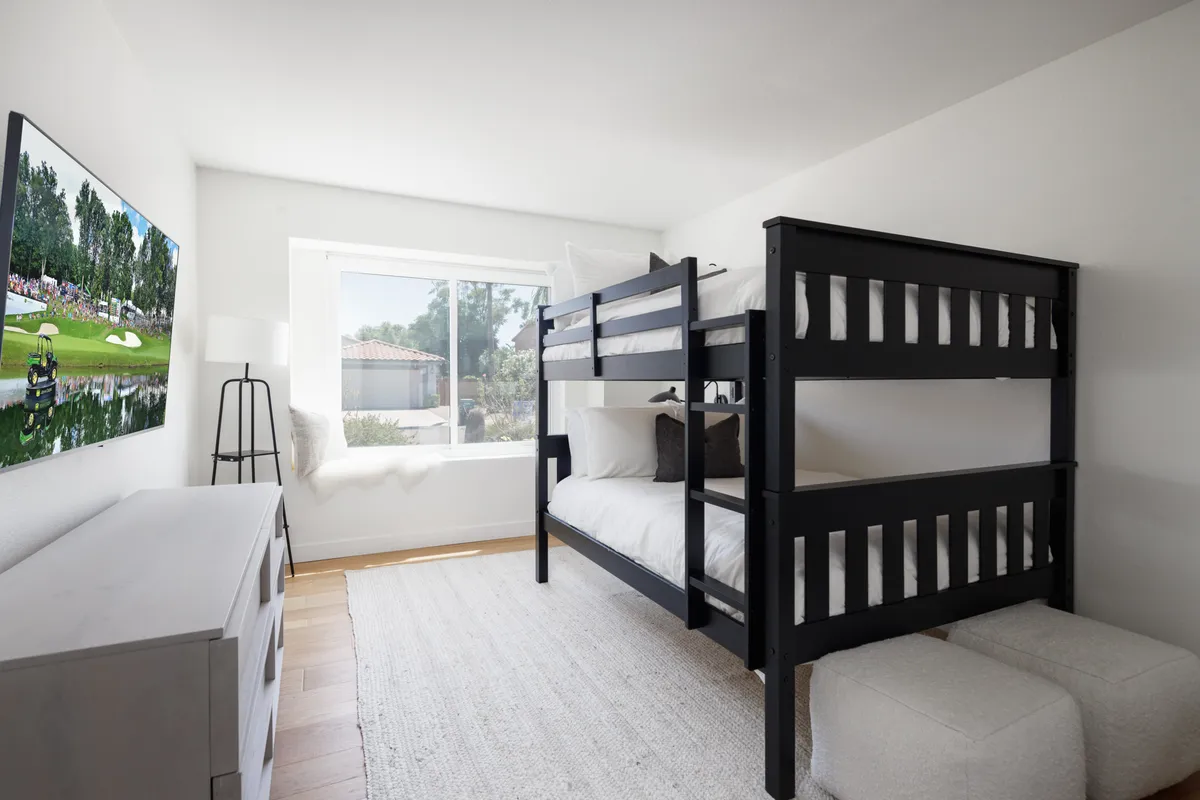 A playful, serene bedroom with a bunk bed, large window for natural light, a television, dresser, and a soft floor rug. The space feels both inviting and practical for families with children.