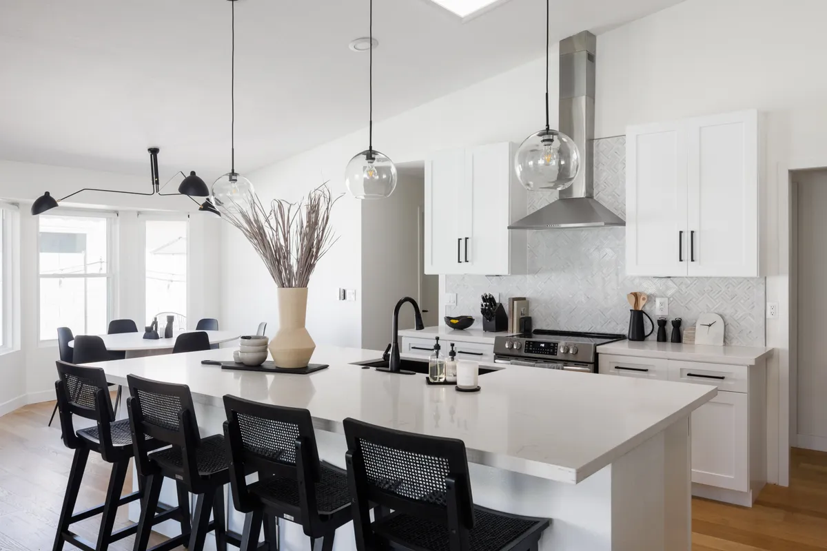 A contemporary kitchen featuring a spacious island with pendant lights, a vase with dried plants, and stainless steel appliances. The space is bright, welcoming, and set up for both meal prep and social interaction, reflecting a sense of thoughtful hospitality and readiness for new arrivals.