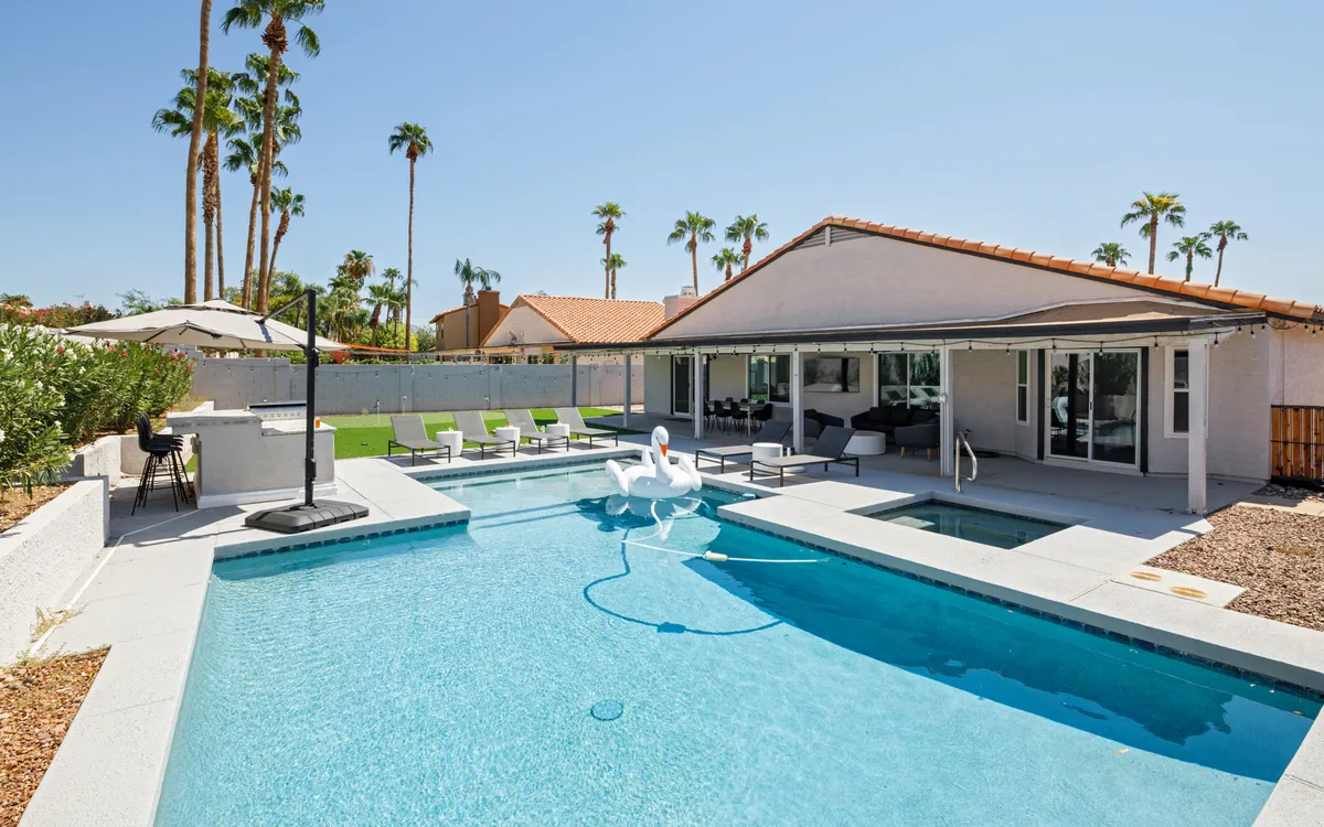 This inviting exterior shot captures the sparkling swimming pool with a playful swan float, surrounded by an outdoor seating area and framed by palm trees. The scene is filled with sunlight and exudes a cheerful, resort-like atmosphere.