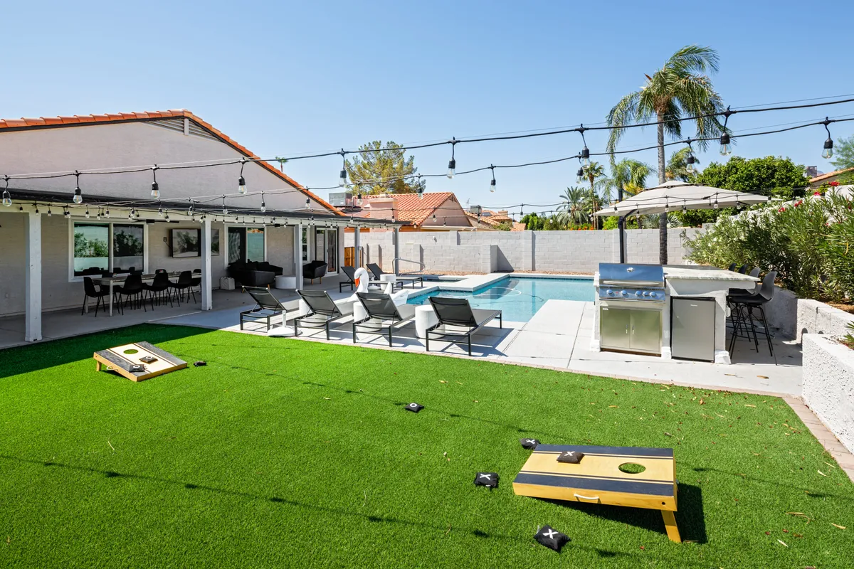 The image showcases a lush backyard oasis at sunset featuring a sparkling swimming pool framed by inviting lounge chairs, a state-of-the-art outdoor grill, festive string lights overhead, and a cornhole game set up beside the pool. The scene radiates a relaxed, social vibe, perfect for gatherings and fun under the Arizona sky.