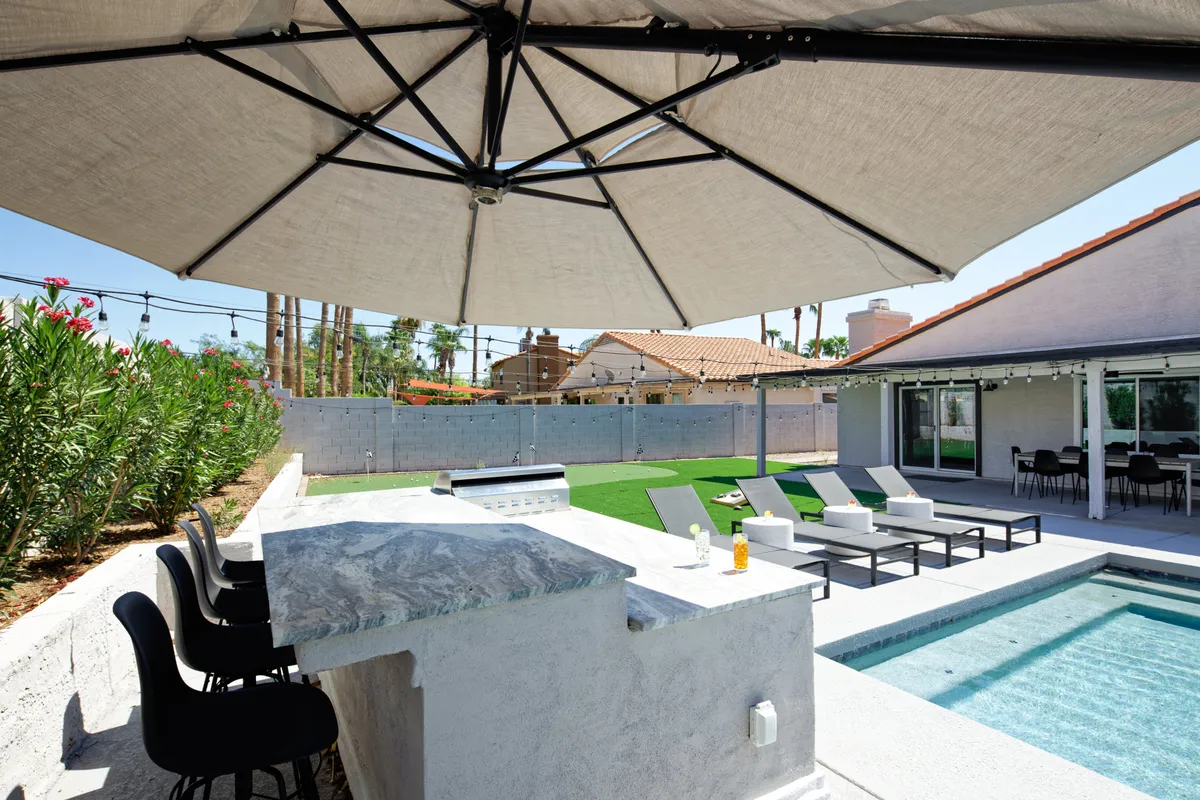 This exterior image highlights an outdoor bar area adjacent to a swimming pool, with comfortable lounge chairs and umbrella shade. Decorative plants soften the modern style, and the area looks set for both quiet, romantic coffee mornings and lively poolside afternoons.