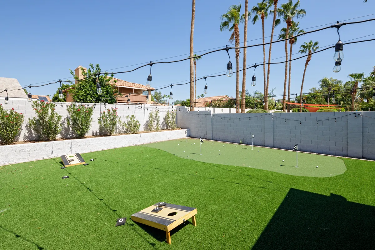 A wide-angle exterior view displays the artificial grass lawn set up for cornhole, with string lights overhead, a visible putting green, and a backdrop of the yard's inviting amenities. The playful setup encourages outdoor games and gatherings.