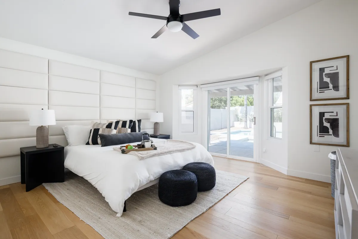 A serene and modern bedroom featuring a king bed adorned with plush decorative pillows and nightstands with lamps. The room opens to the outdoors through sliding glass doors, flooding the space with natural light, and there is a cozy nook ideal for quiet relaxation. Chic, understated artwork completes the restful atmosphere.