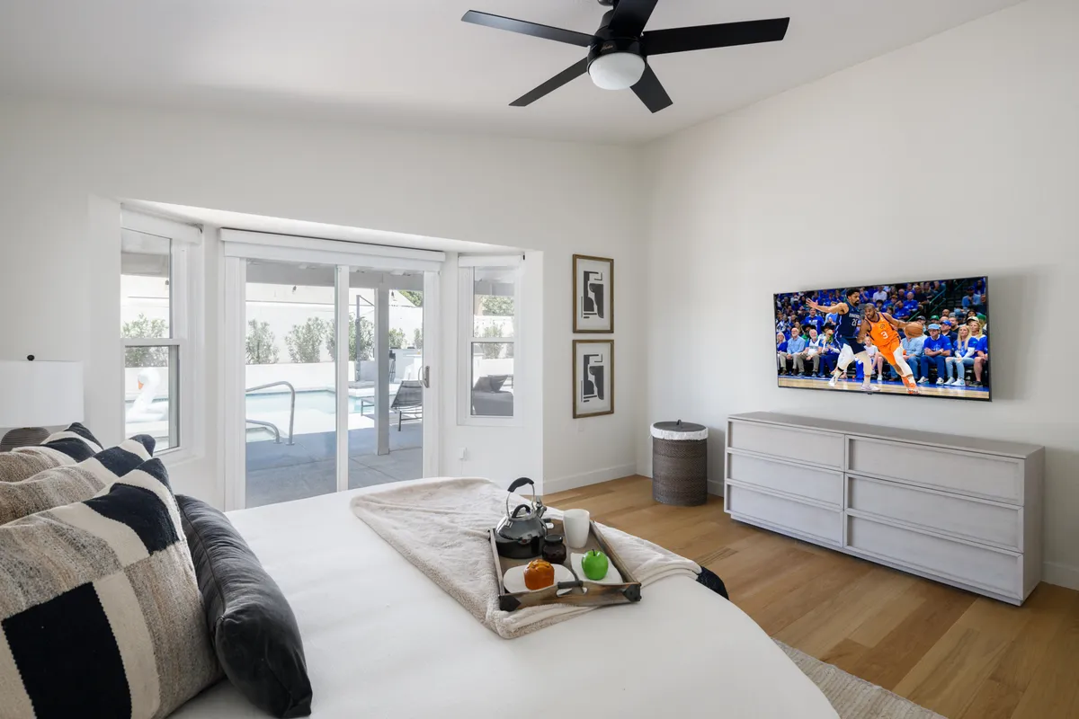 A comfortable bedroom featuring a queen bed with decorative pillows, a television displaying a sports game, and sliding doors that lead directly to the pool area. The space is adorned with contemporary artwork and offers a cozy yet energetic mood, perfect for relaxing or winding down after a day of activities.